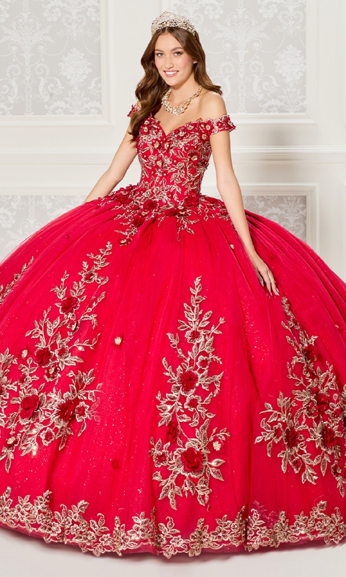 A woman in the Quinceanera Dress PR30114 By Princesa—bright red, off-the-shoulder with gold 3D floral appliques—wears a tiara and jeweled necklace as she stands in an ornate white room.